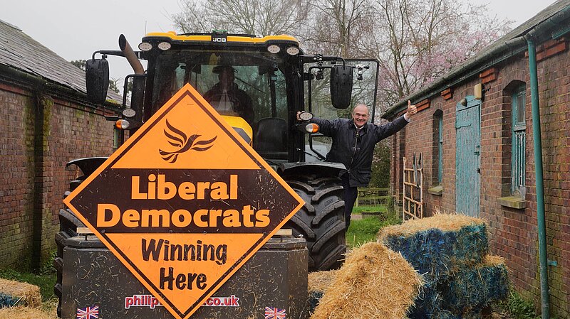 Ed Davey waves from a tractor with a Lib Dem Diamond sign, having knocked down a blue wall of bales