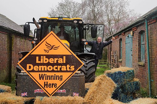 Ed Davey waves from a tractor with a Lib Dem Diamond sign, having knocked down a blue wall of bales